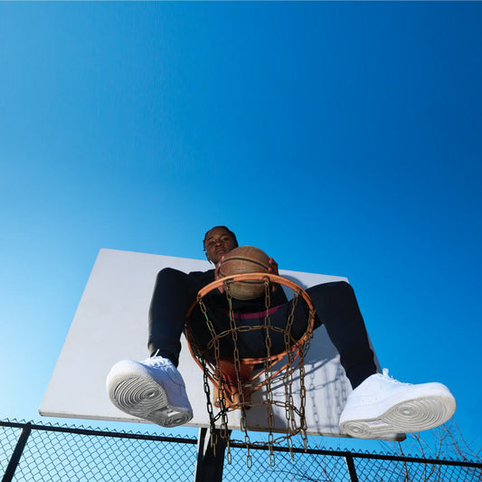 Basketball Spieler in schwarzer Jeans und T-Shirt sitzt bei blauem Himmel im Korb mit Basketball in der Hand und posiert für den Roger Rockawoo Blog Artikel Basketball 3x3