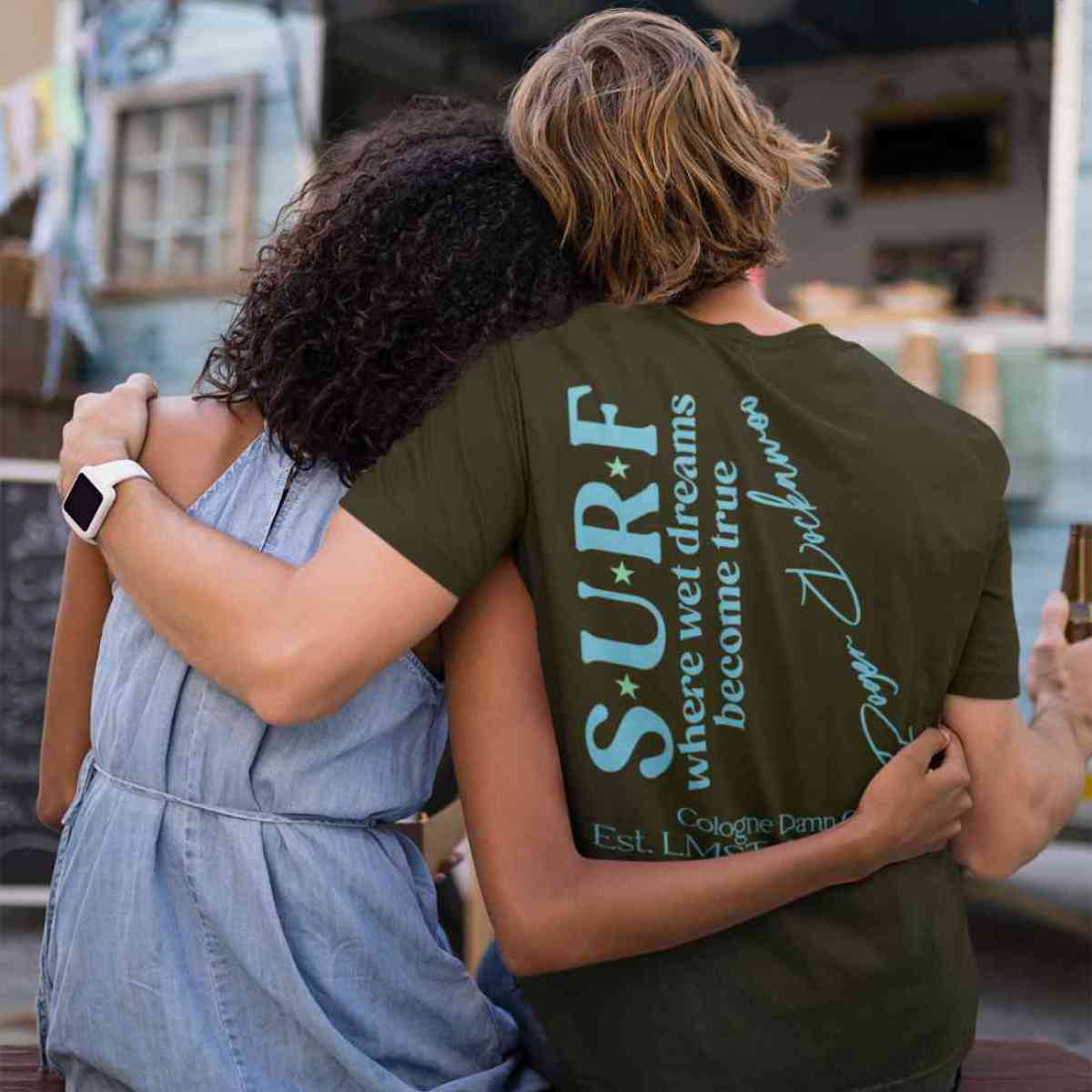 Khaki T-Shirt aus der Surf where wet dreams become true Kollektion von Roger Rockawoo Clothing präsentiert von einem männlichen Model das in der Rückansicht arm in arm mit einer Freundin vor einer Strandhütte sitzt