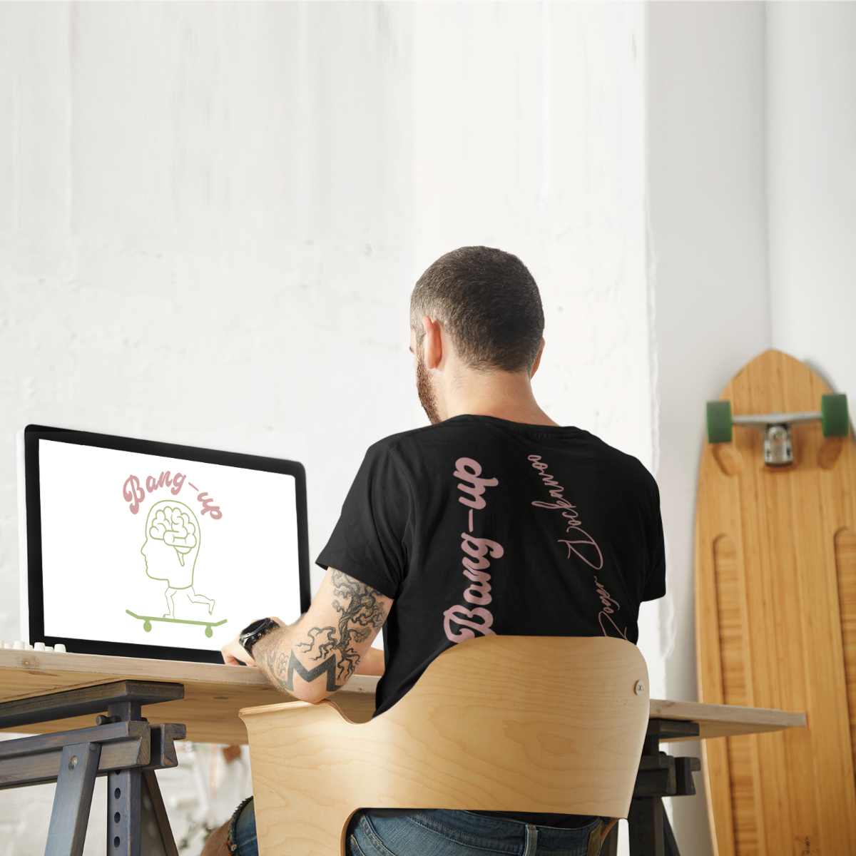 Schwarzes T-Shirt aus der Skateboard bang up brain Kollektion von Roger Rockawoo Clothing präsentiert von einem männlichen Models das am Schreibtisch sitz in der Rückansicht mit Longboard an der Wand