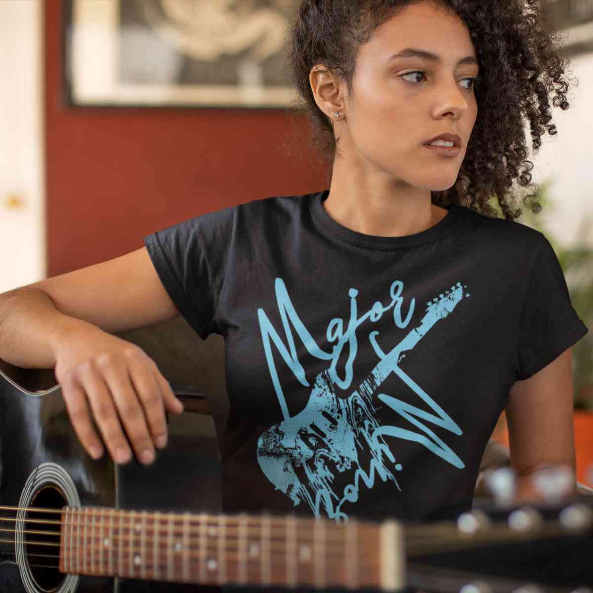 Schwarzes T-Shirt aus der Guitar Major or Minor Kollektion von Roger Rockawoo Clothing präsentiert von weiblichem Model das mit einer schwarzen Akustik Gitarre sitzt und zur Seite schaut