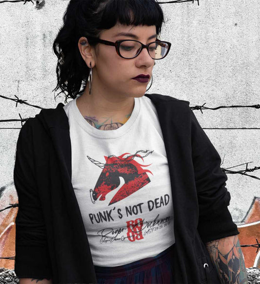 Weibliches junges Model mit schwarzen Haaren und Brille steht vor Graffiti Wand und trägt ein weisses T-Shirt das bedruckt ist mit dem Punker Einhorn aus der Punks not Dead Kollektion von Roger Rockawoo